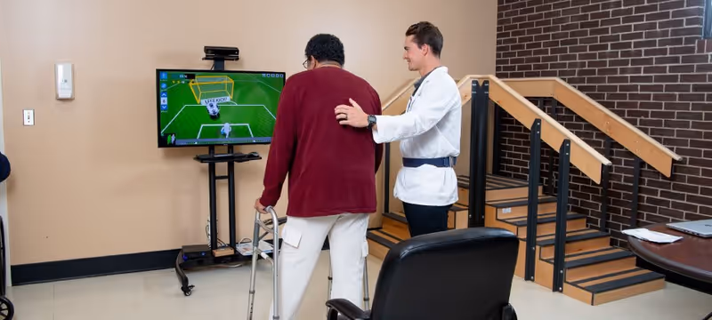 A healthcare professional assists an elderly person using a walker in a rehabilitation room. They are facing a television screen displaying a virtual soccer game. The room has a small set of stairs with handrails and a table with a laptop on it.