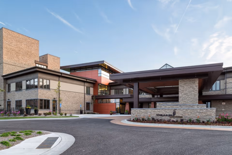 Exterior view of a modern senior living facility building with a covered entrance, stone and brick facade, large windows, and landscaped surroundings under a clear sky.