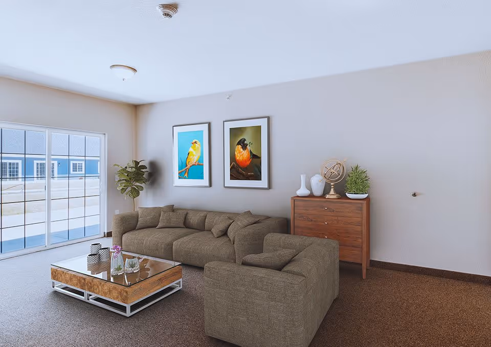 A cozy living room with a large brown sectional sofa and a matching armchair. A wooden coffee table with a glass top is in front of the sofa, decorated with small plants and a flower vase. On the wall above a wooden chest of drawers are two framed pictures of colorful birds. A potted plant is placed in the corner near a large sliding glass door that lets in natural light.