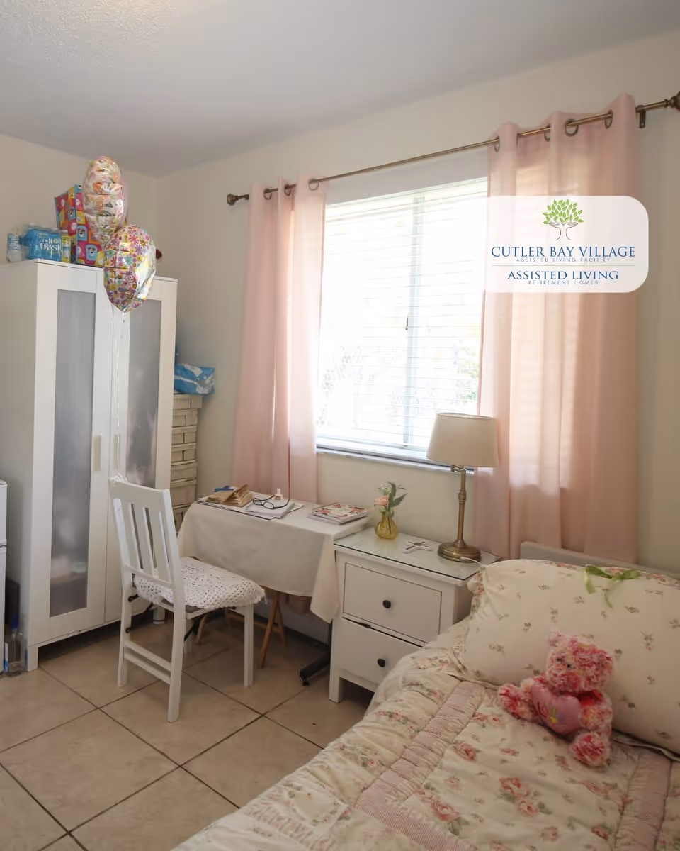 A cozy bedroom with a floral-patterned bedspread and a pink teddy bear on the bed. Next to the bed is a white nightstand with a small lamp and a vase with flowers. A small table covered with a white cloth holds some books and glasses, accompanied by a white chair with a crocheted seat cover. A white wardrobe stands in the corner with balloons attached to it. The window is covered with pink curtains, allowing natural light to brighten the room.