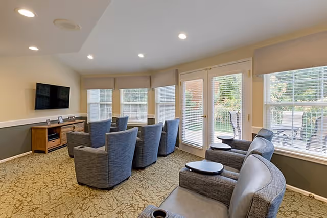 Cozy common room with several gray swivel armchairs facing a wall-mounted TV and large windows and a glass door to an outdoor patio.