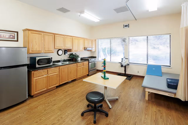 A room with wooden cabinets, a stainless steel refrigerator, microwave, stove, and sink along one wall. The room has large windows with blinds, a small table with a black rolling stool, and a therapy or examination table with a blue cushion. The floor is wood, and there is a fan mounted near the windows.
