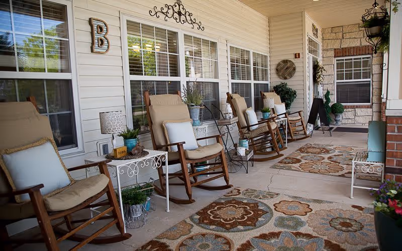 A covered outdoor porch area with several cushioned wooden rocking chairs arranged along the wall of a building. The porch has decorative rugs with floral patterns on the floor, small side tables with plants and lamps, and wall decorations including a large letter 'B' and a metal wall art piece. The area appears inviting and comfortable for sitting and relaxing.