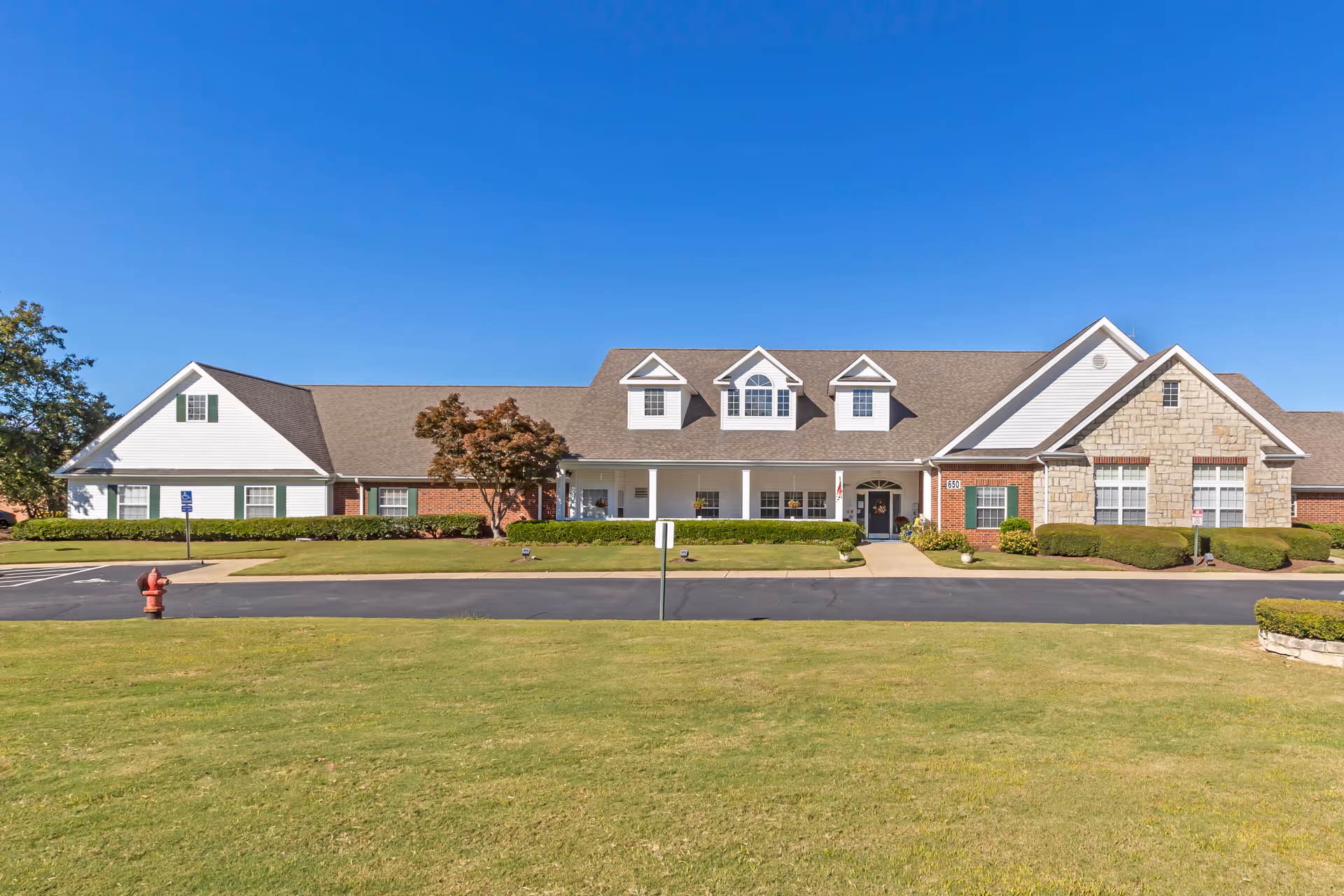 Front exterior view of a single-story senior living facility building with a combination of brick, stone, and white siding. The building features a covered entrance with a walkway, several windows, and dormer windows on the roof. The foreground shows a well-maintained lawn, a fire hydrant, and a paved driveway under a clear blue sky.
