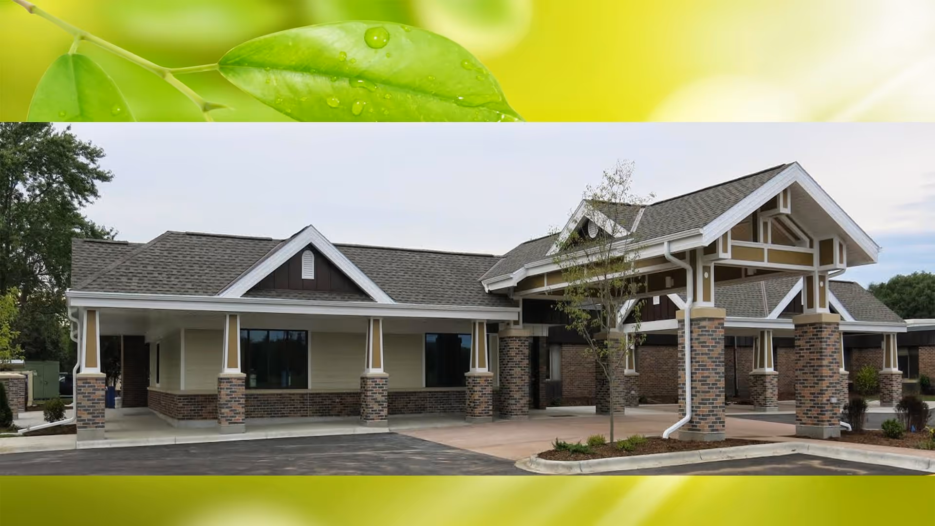 Exterior view of a single-story senior living facility building with a covered entrance supported by brick columns, a gray shingled roof, and beige siding. There is a small tree and landscaped area near the entrance, and the sky is partly cloudy.