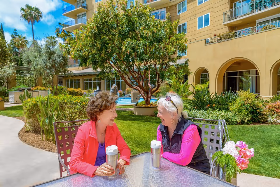 Two elderly women sitting at a glass outdoor table in a garden courtyard of a senior living facility, smiling and enjoying conversation. The background shows green grass, trees, shrubs, and a multi-story building with balconies and arched windows.