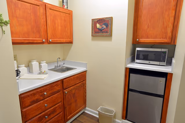Small kitchenette with wooden cabinets, a stainless steel sink, a microwave on a countertop, a mini refrigerator below the microwave, a small trash bin on the floor, and a decorative framed picture on the wall.
