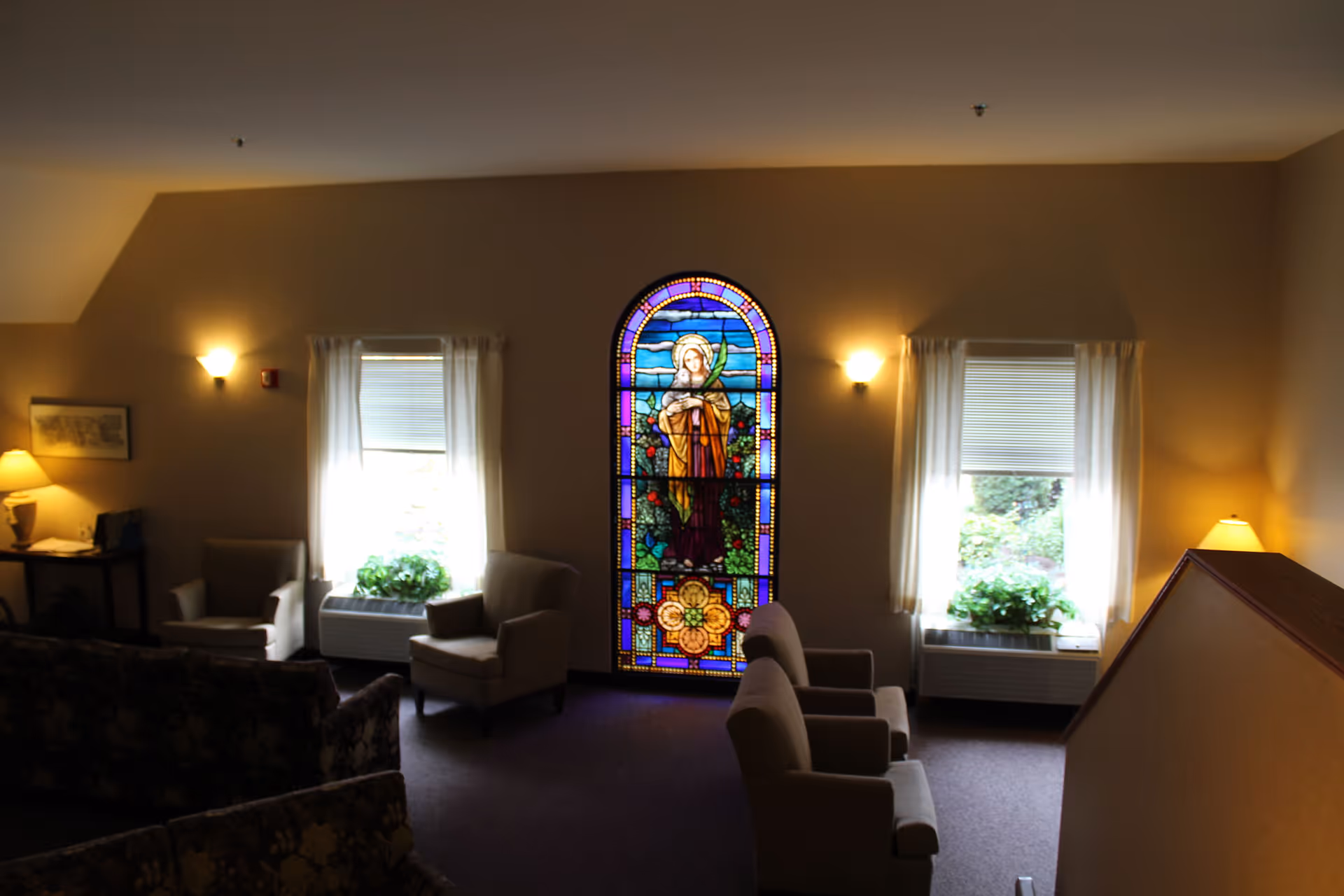Interior view of a lounge area with several armchairs and sofas arranged around a stained glass window depicting a religious figure. The room has two windows with white curtains and plants on the windowsills. Warm lighting is provided by wall sconces and table lamps.