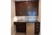 Small kitchenette area with dark wood cabinets, a granite countertop, a small sink, and a mini refrigerator underneath the counter.