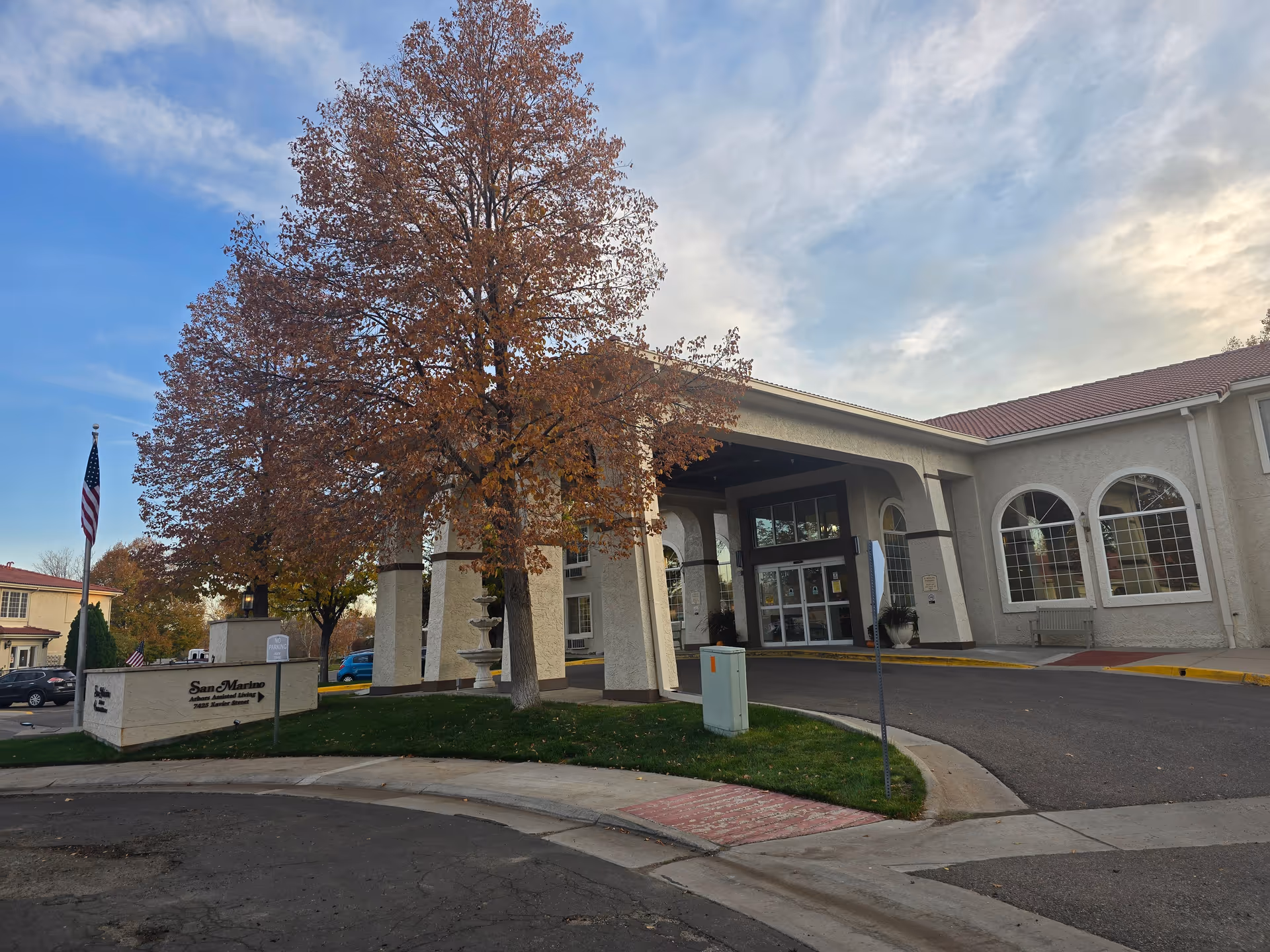 Front entrance of the San Marino Retirement Community building with a covered porte-cochère, a large tree with autumn leaves, and an American flag.