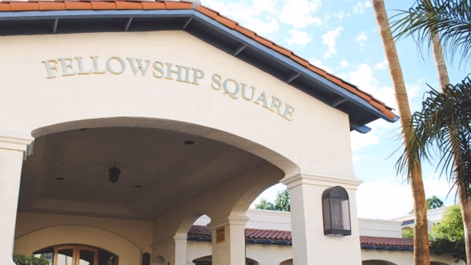 Entrance of Fellowship Square building with a covered archway, beige walls, and a red tile roof. Palm trees and blue sky are visible in the background.