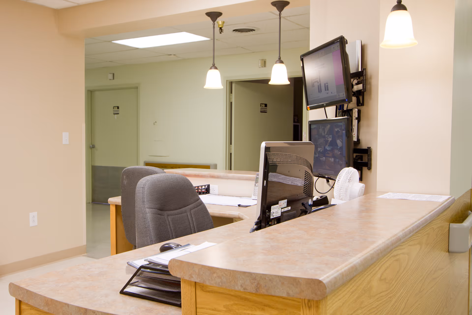 Reception desk area with two computer monitors, office chairs, and hanging pendant lights in a healthcare or senior living facility. Doors and hallway are visible in the background.