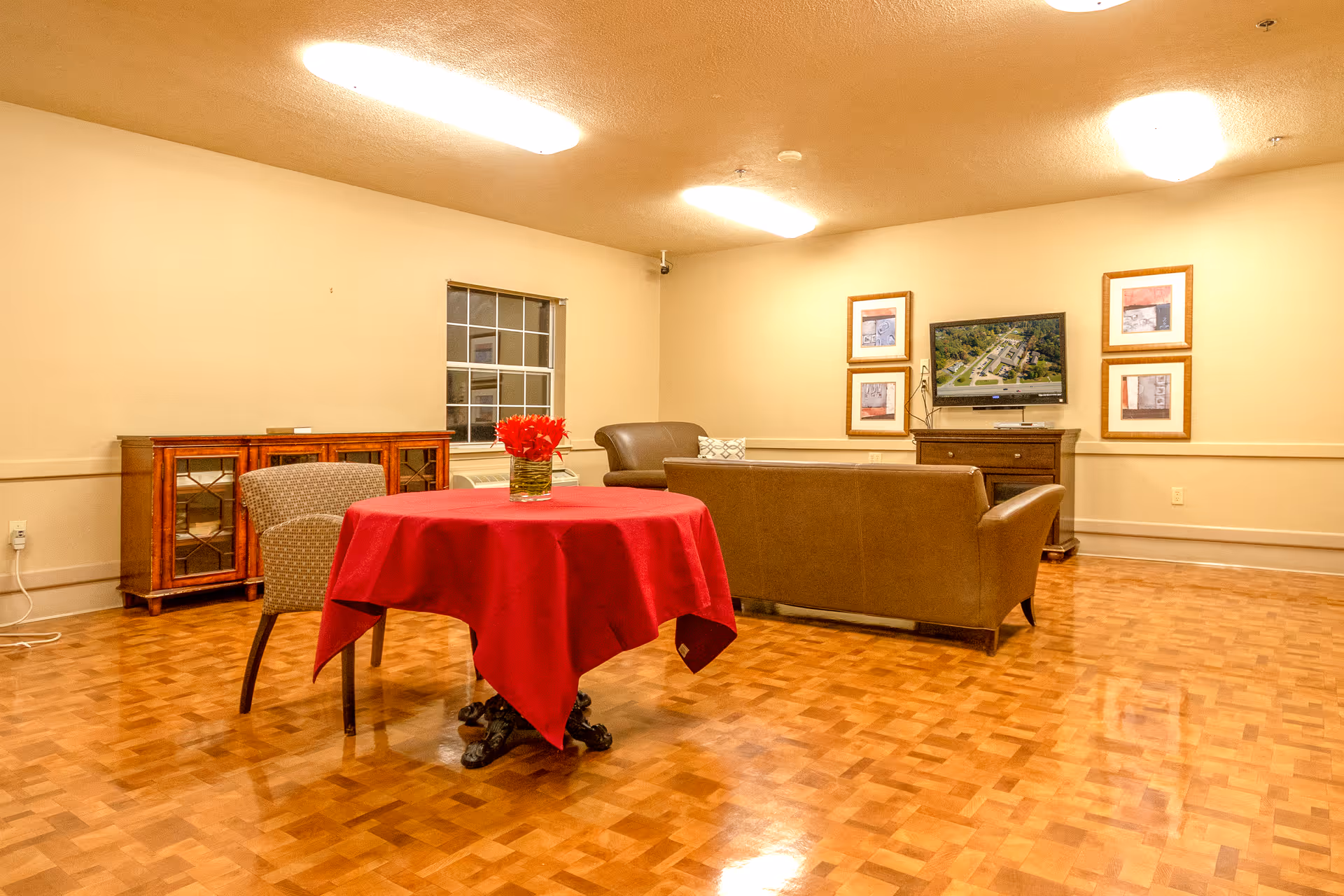 A cozy living room area with a wooden parquet floor, a round table covered with a red tablecloth and a vase of red flowers, two armchairs, a brown leather sofa, a wooden cabinet, a window, and a wall-mounted TV surrounded by framed pictures.