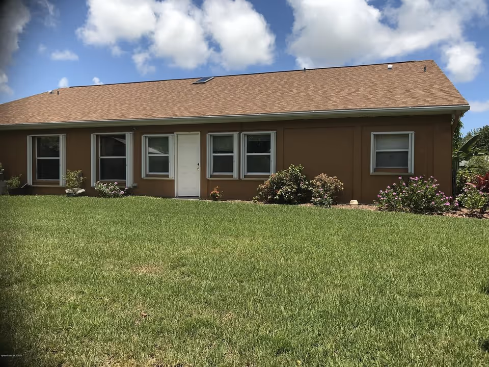 Single-story brown house with a white door, several windows, flowering shrubs, and a grassy front yard under a blue sky.
