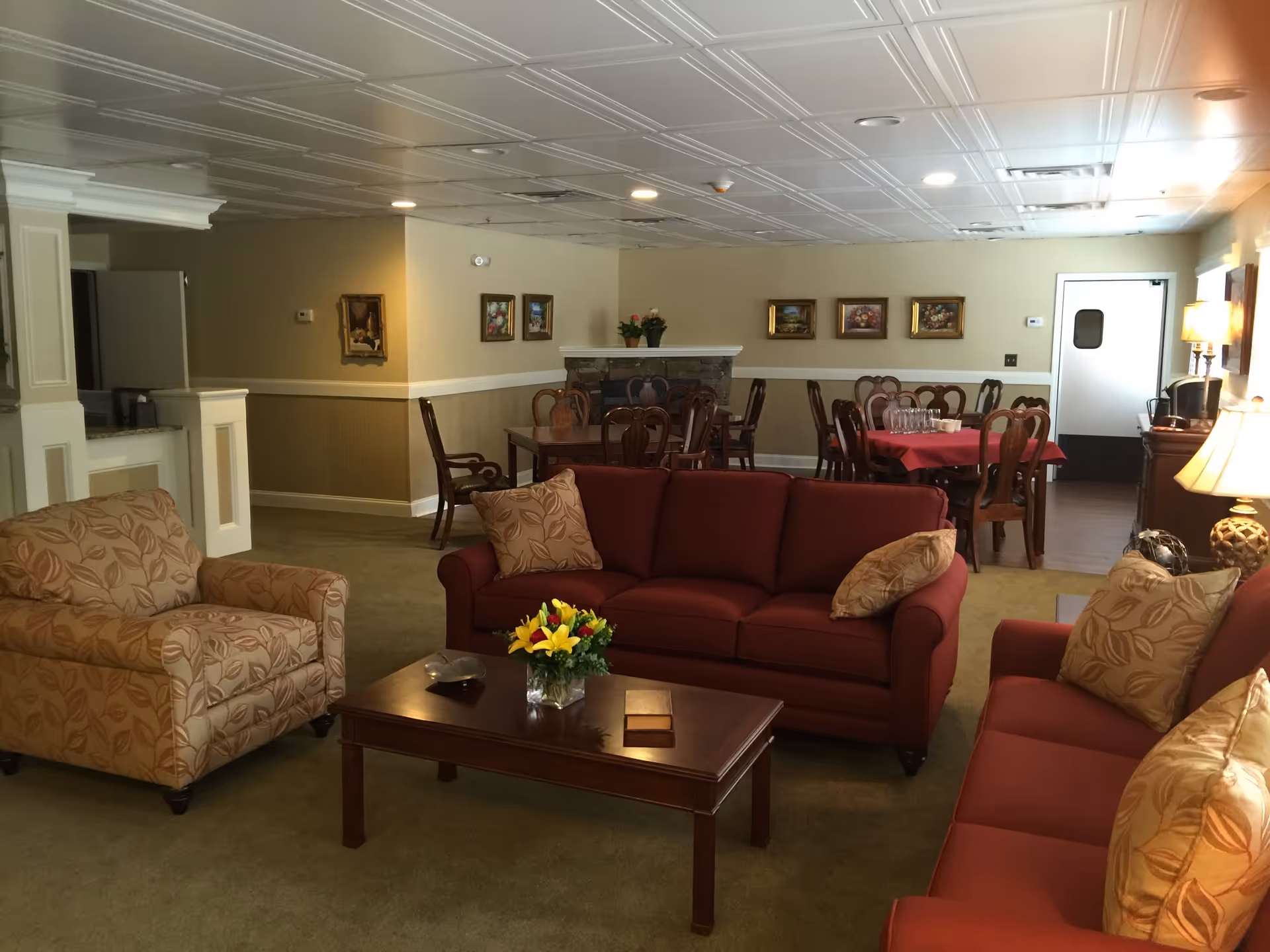 A cozy senior living community common area featuring two red sofas and one beige armchair with patterned cushions arranged around a wooden coffee table with a flower vase. In the background, there are dining tables with chairs, a fireplace, framed paintings on the walls, and warm lighting from lamps.