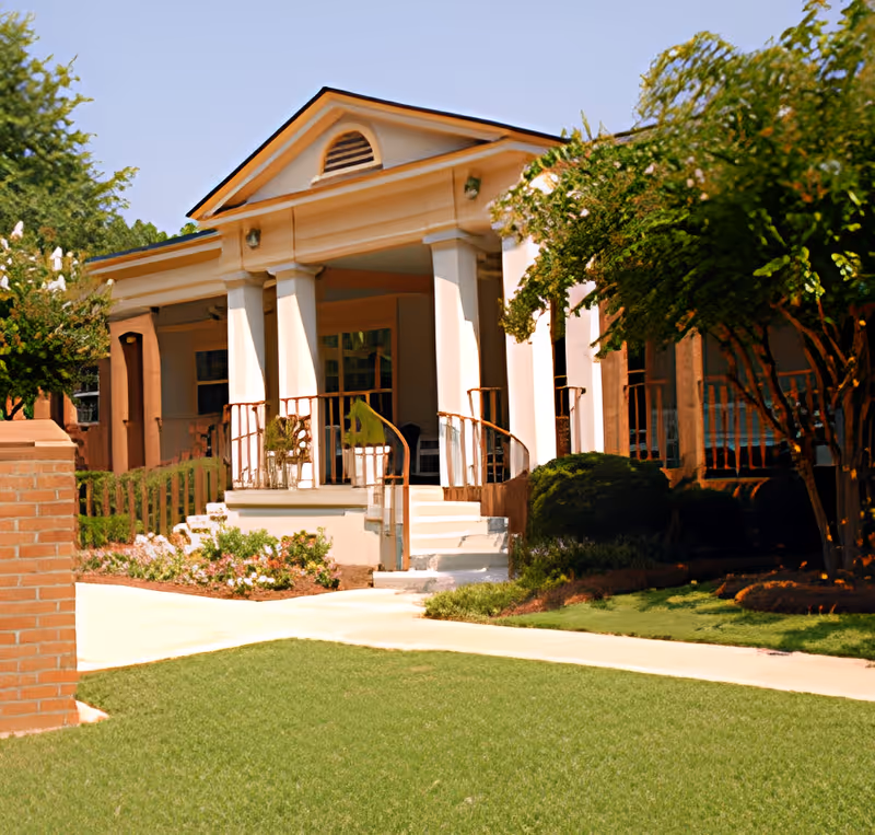 Front entrance of a single-story building with a columned porch, steps and railings, surrounded by landscaped lawn and trees.