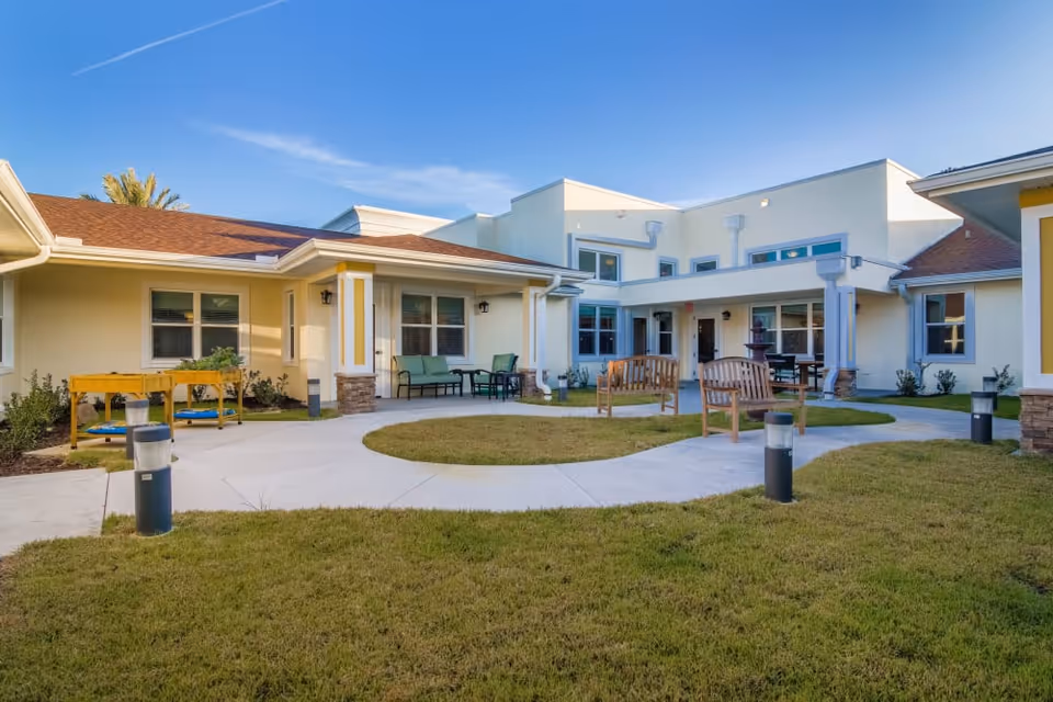 Sunny courtyard with a circular paved walkway, wooden benches, patio seating, and lawn in front of a single-story care facility.