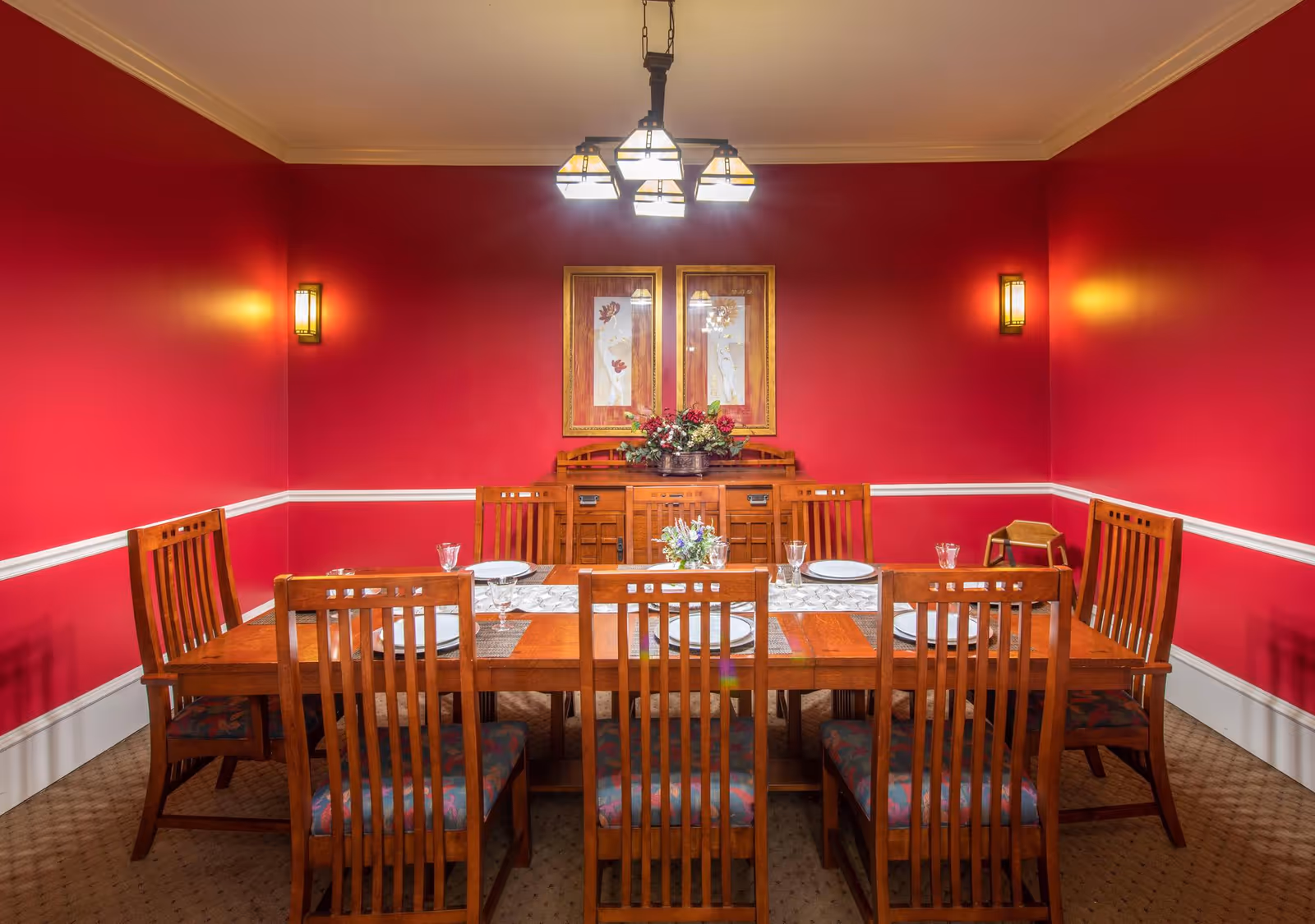 A dining room with bright red walls and white trim, featuring a wooden dining table set for six with plates, glasses, and a floral centerpiece. A wooden sideboard with a flower arrangement and two framed artworks hangs on the wall behind the table. The room is lit by a ceiling light fixture and two wall sconces.