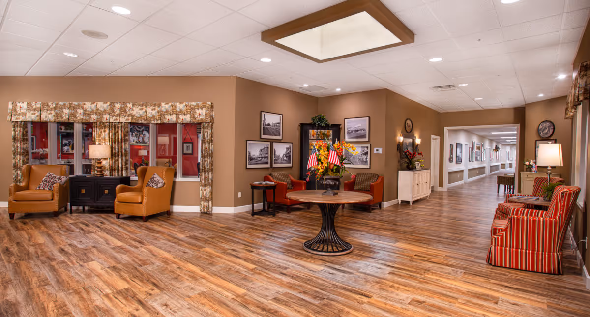Well-lit senior living lobby with armchairs, a central round table topped with flowers and small flags, and a long hallway.