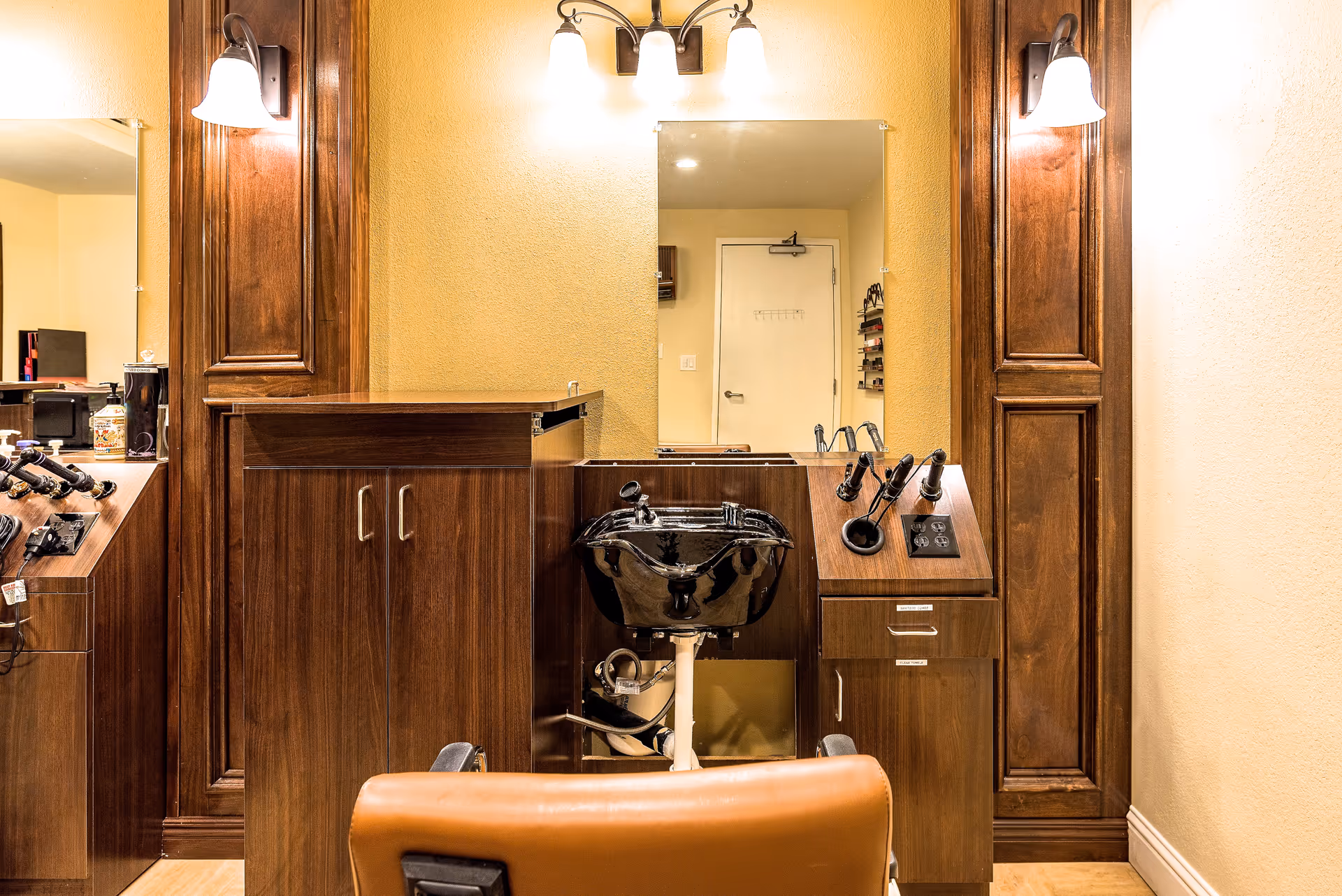 Interior salon station with a black shampoo sink, brown wood cabinetry, mirror, and a salon chair.