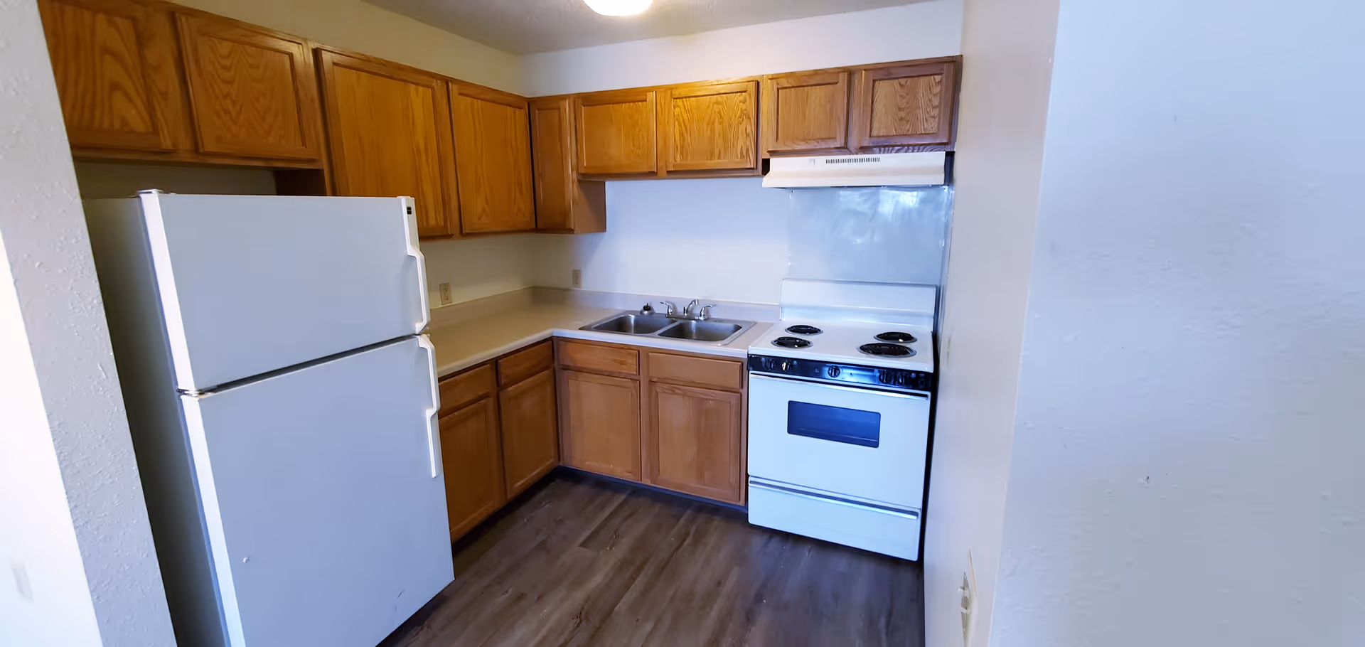 A small kitchen with wooden cabinets, a white refrigerator, a double stainless steel sink, a white electric stove with four burners, and a white range hood. The kitchen has light-colored countertops and wood-look flooring.