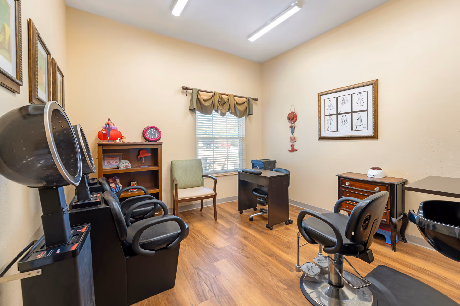 Small salon/styling room with hairdressing chairs, hooded dryers, a desk, shelving and a window in a senior living facility.