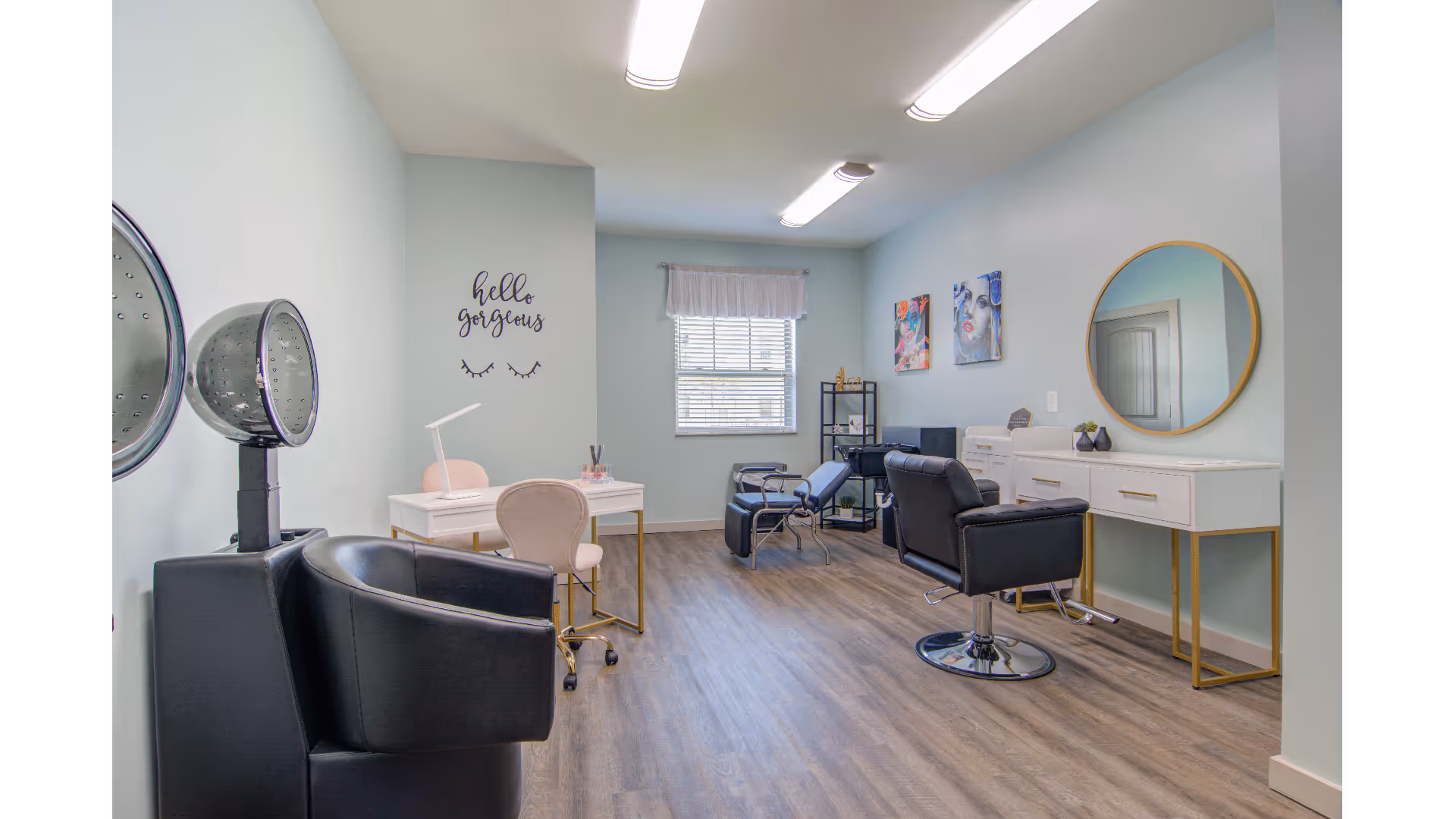 Bright salon-style interior with styling chairs, hooded hair dryers, manicure stations, a round mirror and a 'hello gorgeous' wall decal.
