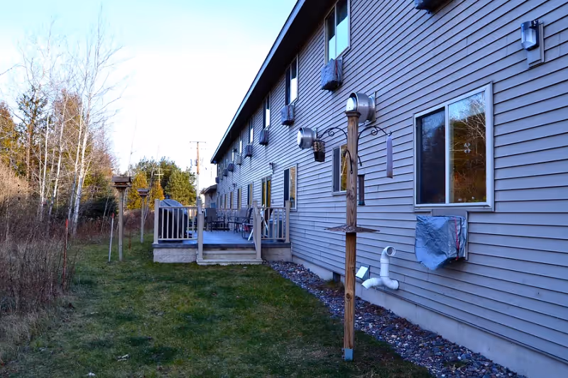 Side exterior of a multi-unit assisted living building with a raised deck, bird feeders on posts, and a grassy yard.