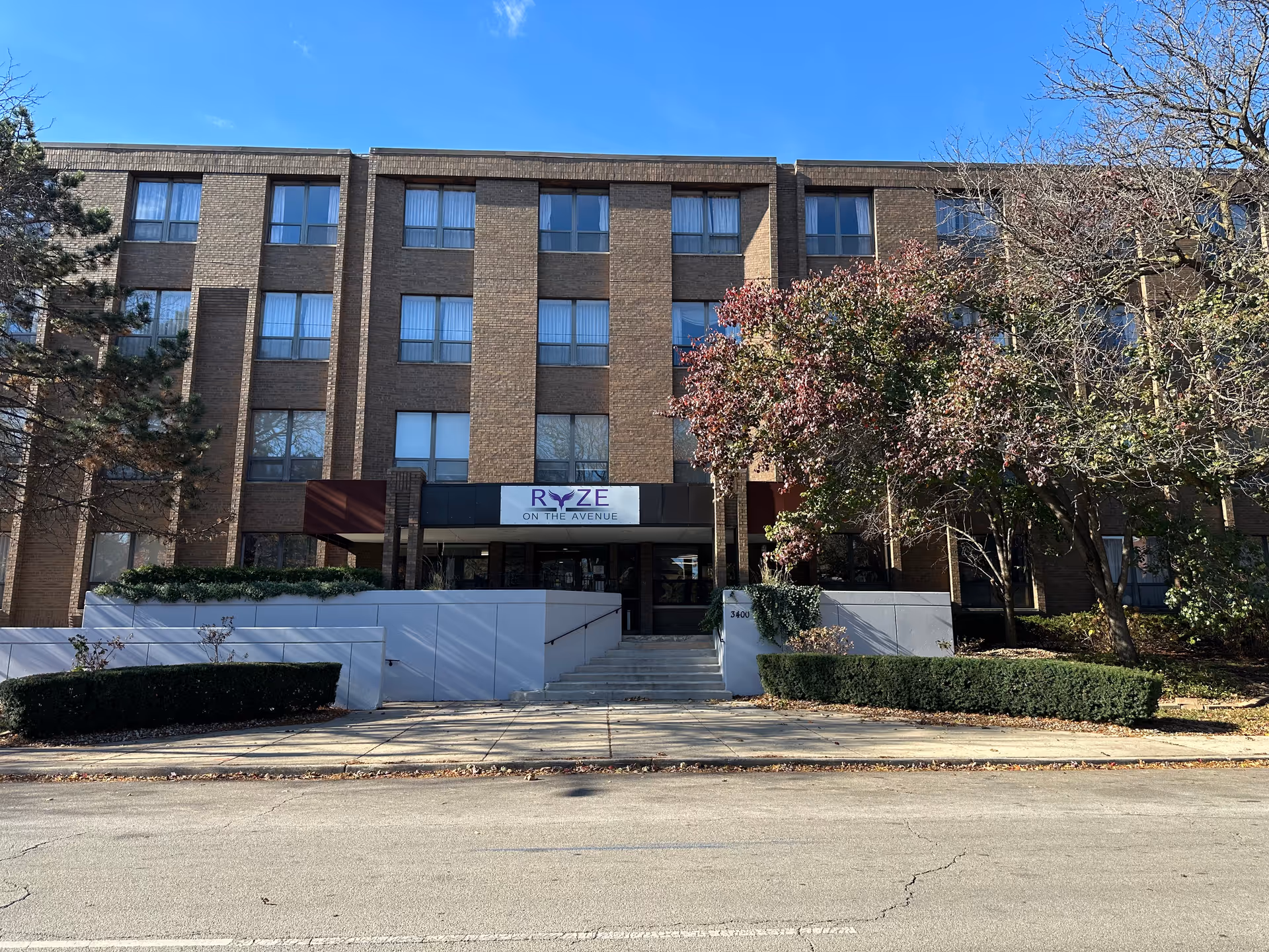 Front exterior of a multi-story brick senior living building with a sign reading 'RYZE on the Avenue', entrance steps, and landscaping.