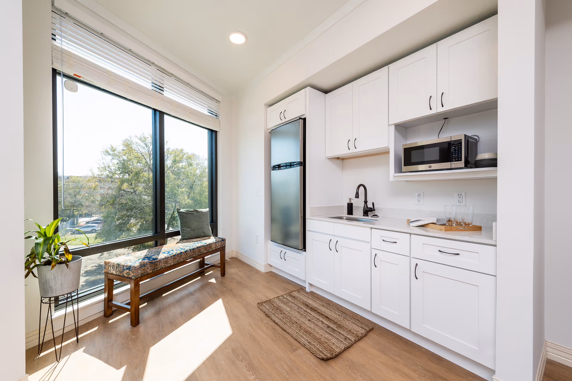 Bright kitchen area with white cabinets, a built-in microwave, a stainless steel refrigerator, and a sink with a black faucet. There is a wooden bench with a cushion and a pillow next to a large window letting in natural light. A small potted plant is on a stand near the window, and a woven rug is on the wooden floor.