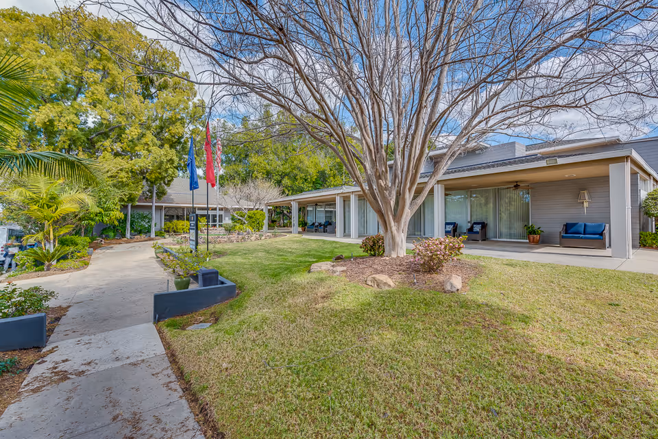 Front exterior of Monte Vista Village with a paved walkway, flags, a large tree, lawn, and a covered patio with seating.