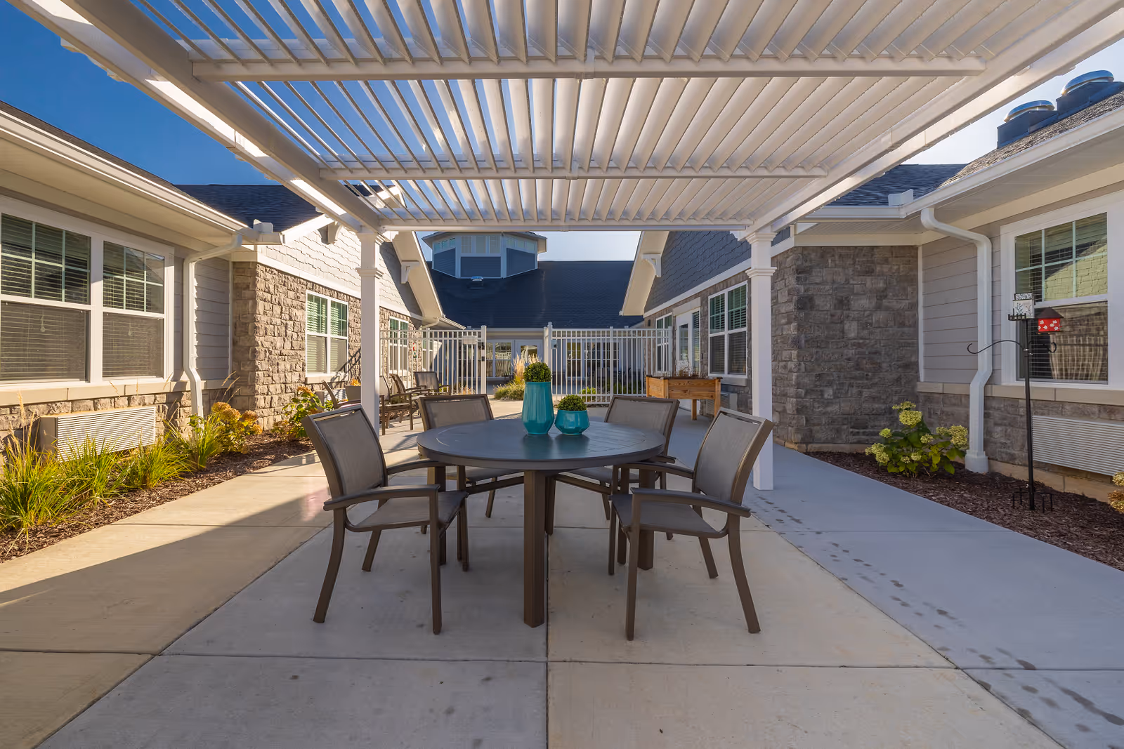 Outdoor patio area at The Springs at Wyandot Trail with a round table and six chairs under a white pergola. The patio is surrounded by single-story buildings with stone and siding exteriors, windows, and landscaped plants along the edges. The sky is clear and blue.