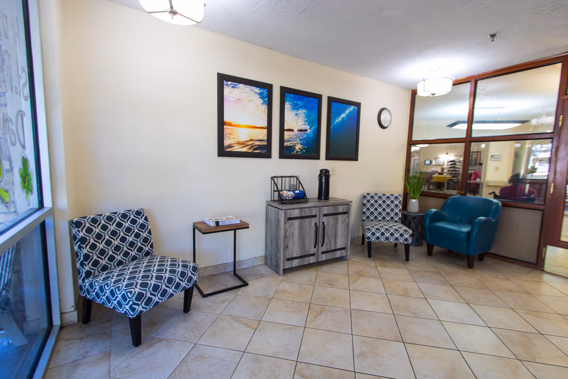 A small waiting area with two patterned chairs, a blue armchair, a small wooden side table, and a wooden cabinet with a coffee dispenser and cups on top. Three framed pictures of ocean scenes hang on the wall above the cabinet. A clock is mounted on the wall, and a large window and glass door are visible.