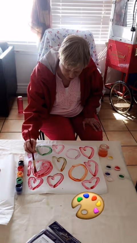 An elderly woman wearing a red jacket and pink shirt is seated at a table painting colorful hearts on a large sheet of paper. Various paint containers and brushes are arranged on the table. The room has tiled floors, a window with blinds, and a red popcorn machine in the background.