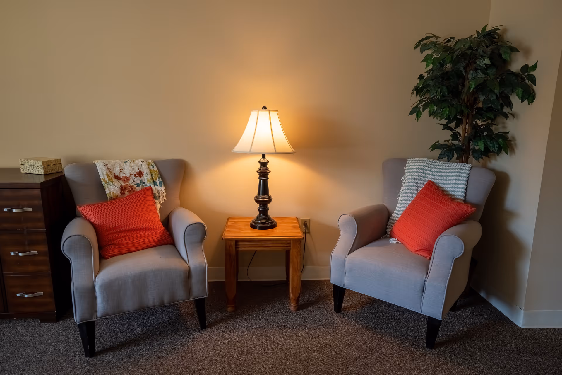 Two gray upholstered armchairs with red pillows and throw blankets are positioned on either side of a wooden side table with a lit table lamp. A wooden chest of drawers is on the left, and a tall green potted plant is on the right against a beige wall.