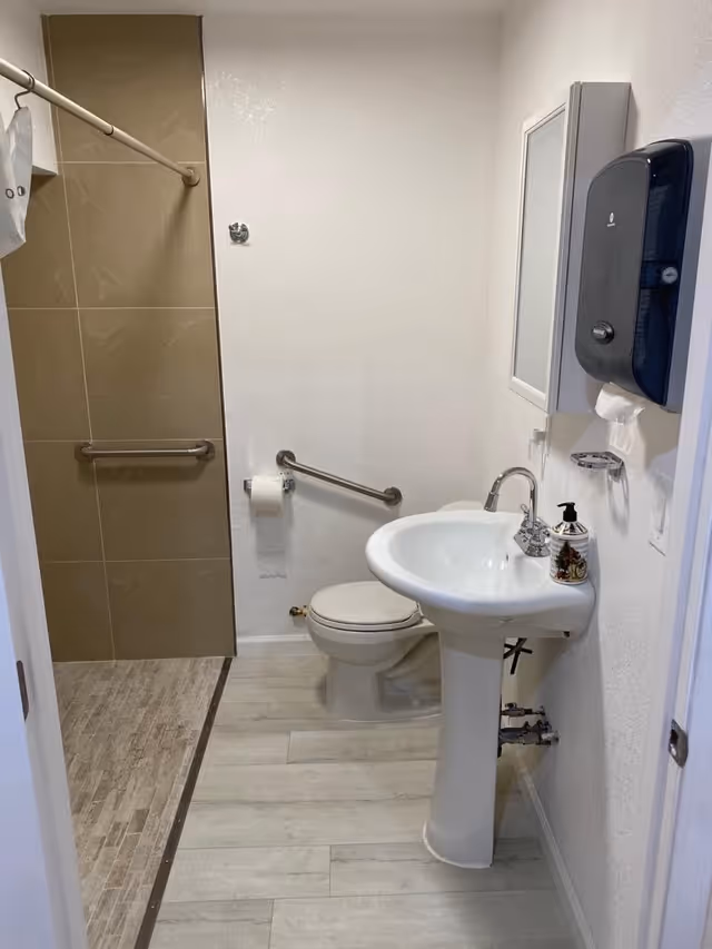 A clean bathroom with a white pedestal sink, a toilet, and a walk-in shower with beige tiles and a grab bar. There is a wall-mounted soap dispenser and a small mirror above the sink. The floor has light-colored wood-like tiles.