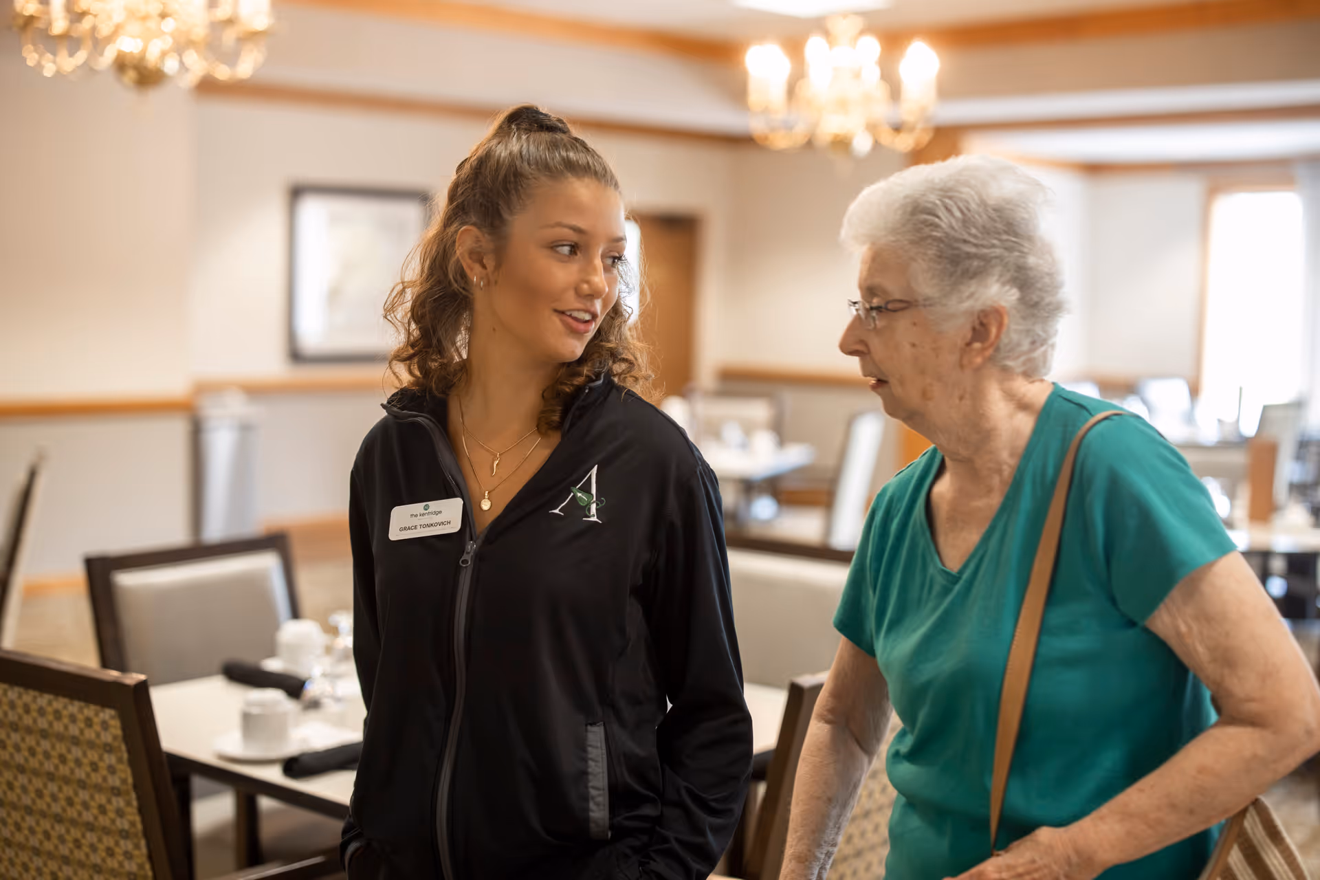A young female staff member wearing a black jacket with a name tag is walking and talking with an elderly woman in a green shirt inside a dining room with tables and chairs set for a meal.