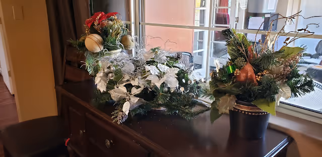A dark wooden sideboard with three festive floral arrangements placed on top, positioned in front of a window with a view of an outdoor patio area. The arrangements include greenery, white and red flowers, and decorative elements like gold ornaments and branches.