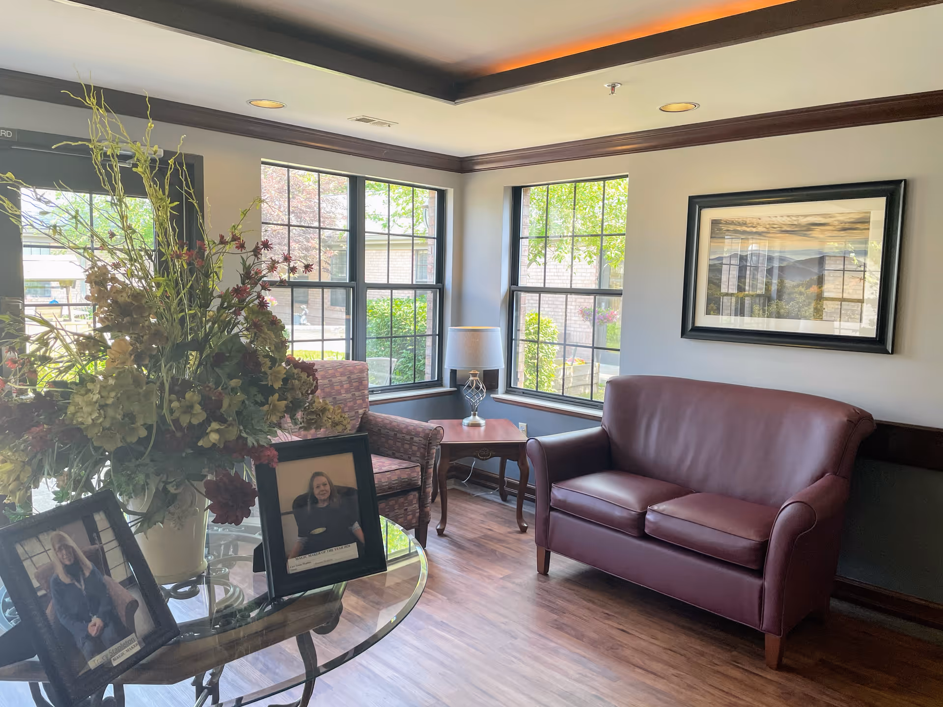 A cozy sitting area in a nursing and rehabilitation facility with a maroon leather loveseat, a patterned armchair, a wooden side table with a lamp, and large windows letting in natural light. A glass table in the foreground holds a large floral arrangement and two framed photographs of staff members. A framed landscape picture hangs on the wall.