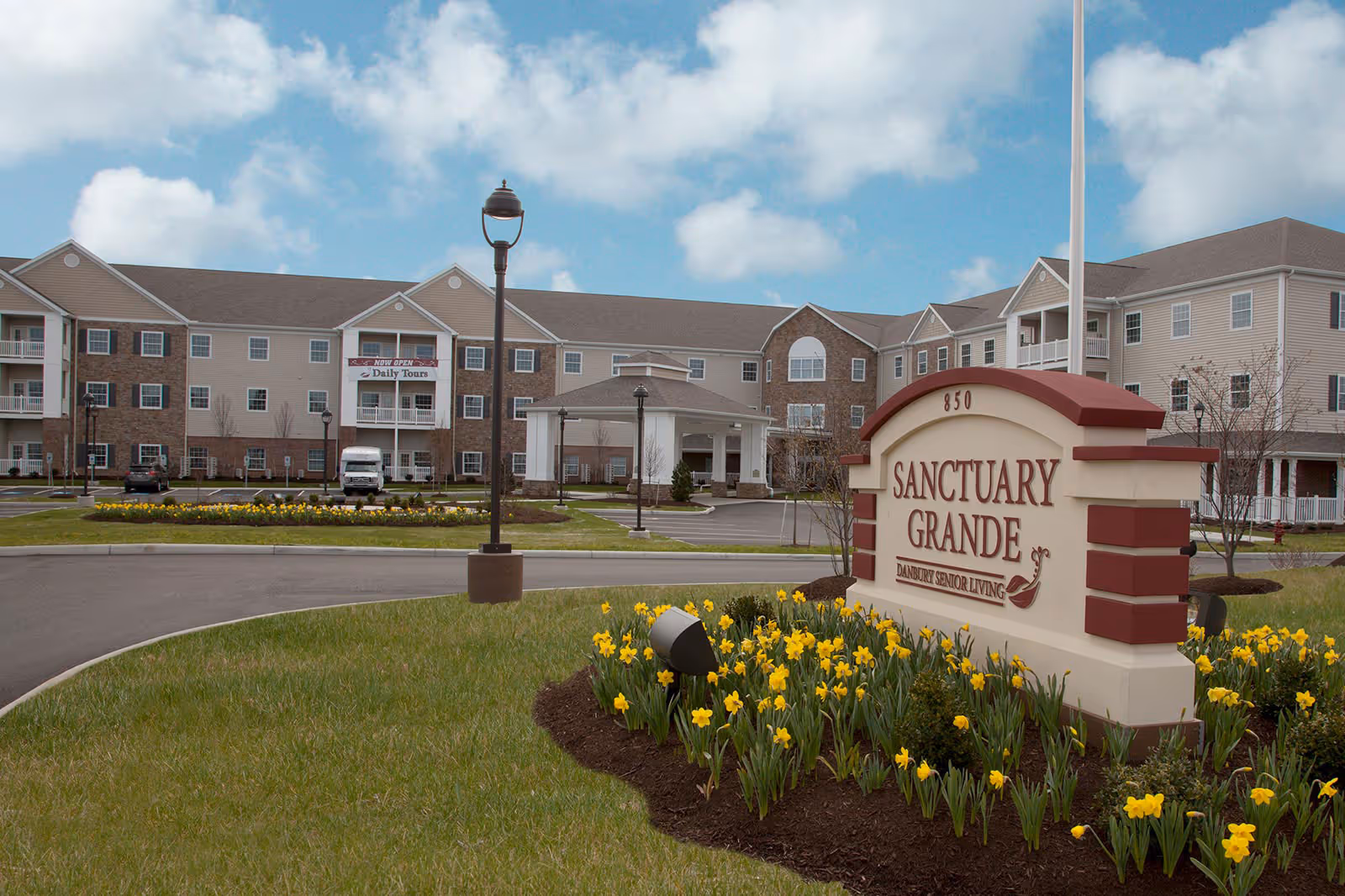 Front exterior of the Sanctuary Grande senior living building with driveway, entrance canopy, and a landscaped sign surrounded by daffodils.