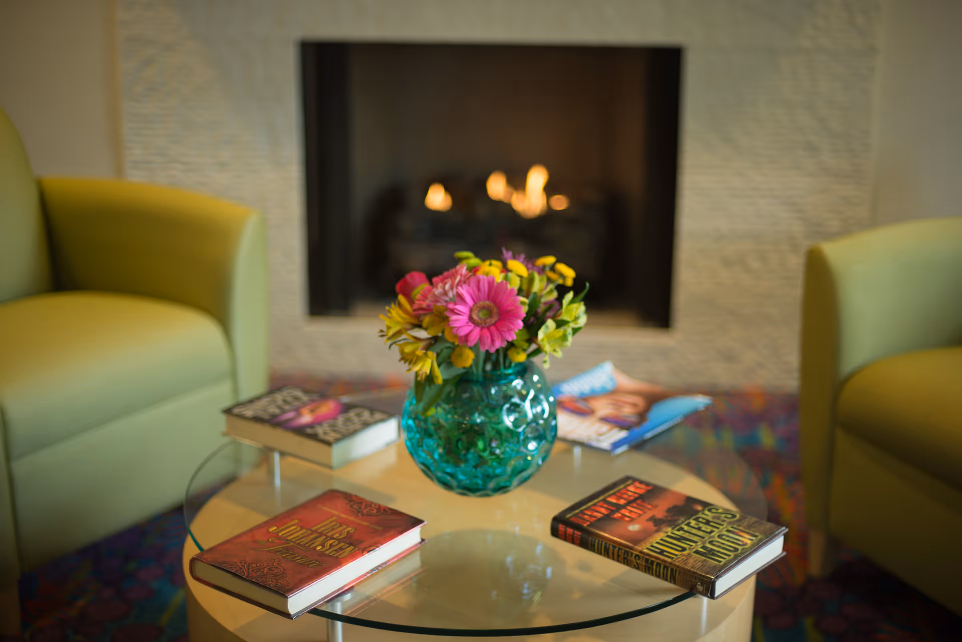 Glass coffee table with a green vase of flowers and several books between two green armchairs in front of a lit fireplace.