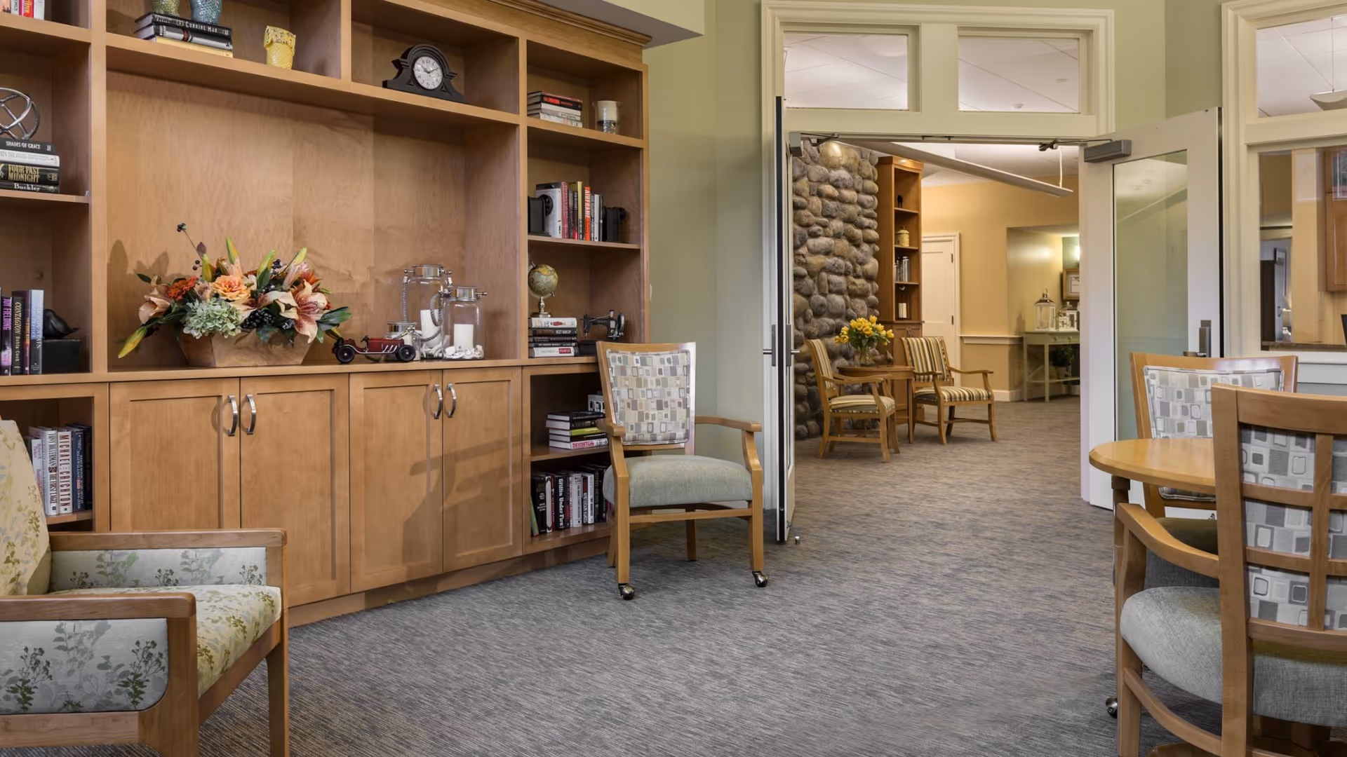 A cozy senior living common room with wooden built-in bookshelves, upholstered chairs, and round tables opening into another seating area.