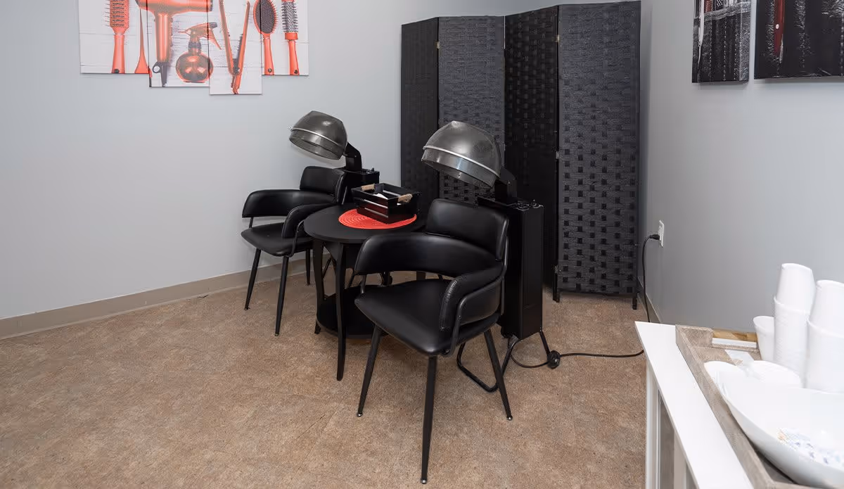 A small salon area with two black salon chairs equipped with hair dryers. There is a round black table between the chairs with a red placemat and a small box on it. A black folding privacy screen is positioned behind the chairs. The walls are light gray with framed pictures of hair styling tools. On the right side, there is a white table with a tray holding stacked white disposable cups and a bowl with small packets.