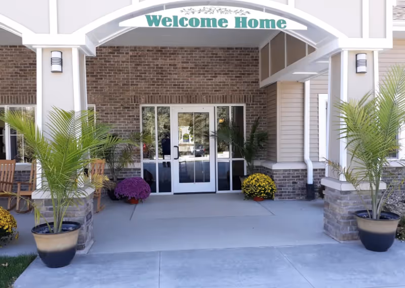 Entrance of a building with a covered porch featuring a 'Welcome Home' sign above the doorway. The entrance has glass double doors flanked by windows, with potted plants and flowers on either side. There are also wooden rocking chairs visible on the left side of the porch.