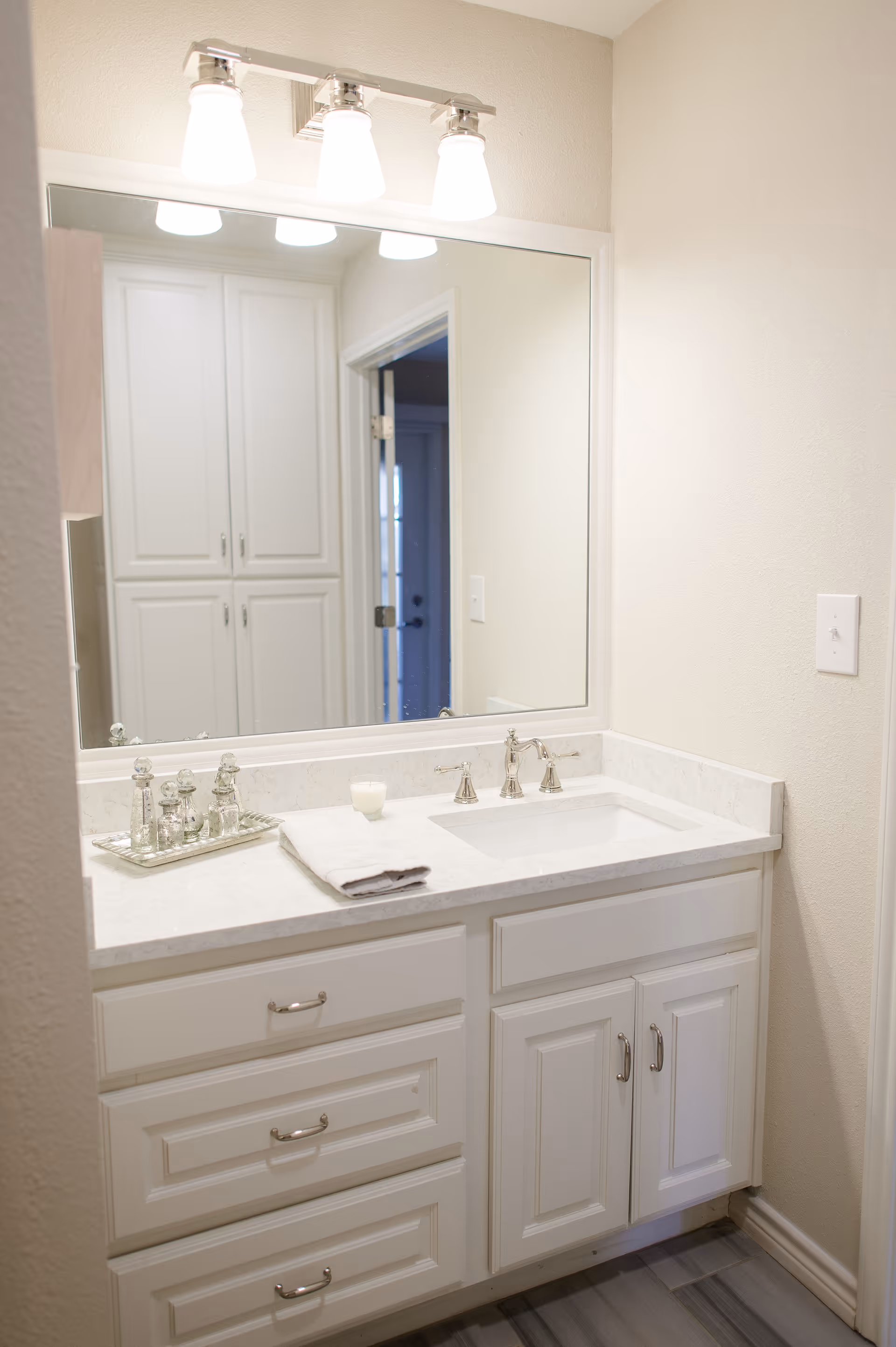 White bathroom vanity with marble countertop, undermount sink, large mirror, and three-light fixture.