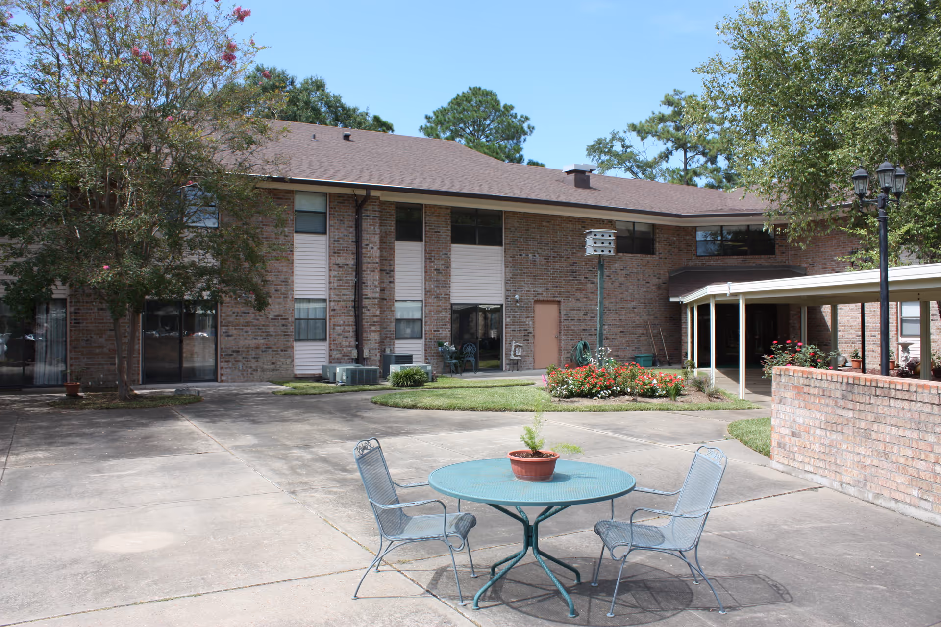 Outdoor courtyard area of a brick building with a round metal table and two metal chairs. There is a potted plant on the table, a flower bed with red flowers, trees, and a covered walkway on the right side. The sky is clear and blue.