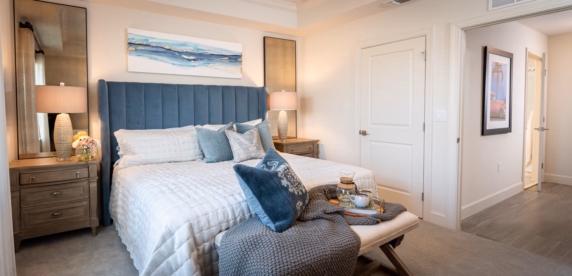 Well-lit furnished bedroom with a blue upholstered headboard, bedside tables and lamps, decorative pillows and a bench holding a tray.