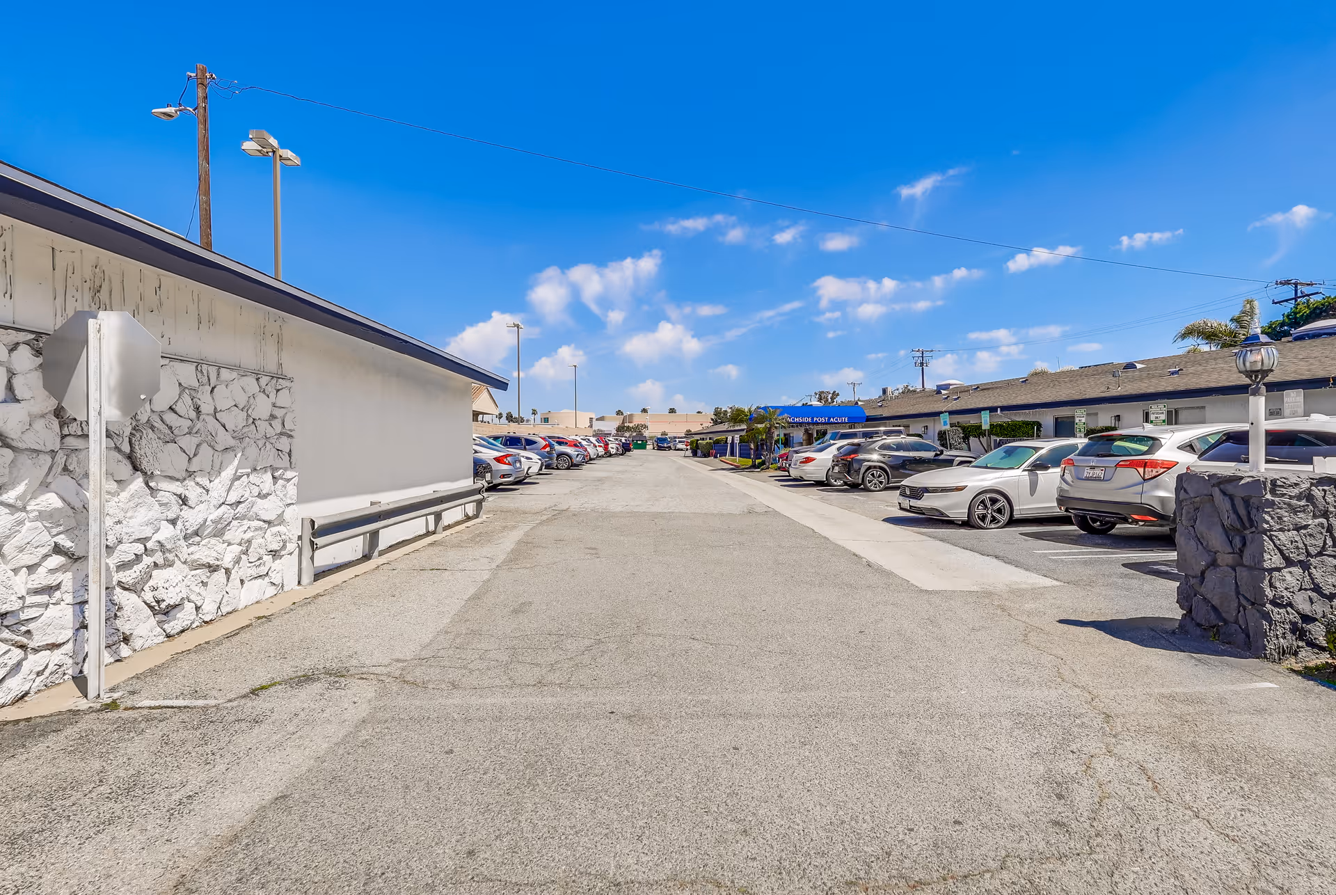 View of a parking lot at Beachside Post Acute facility with cars parked on both sides under a clear blue sky with some clouds. The building is single-story with a stone and stucco exterior.