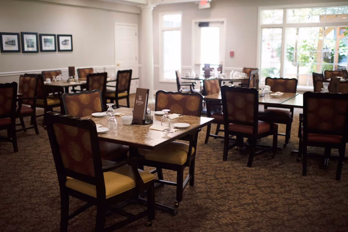 A dining room with multiple tables and chairs arranged neatly. Each table is set with plates, bowls, and upside-down glasses. The room has carpeted flooring, light-colored walls with framed pictures, and large windows allowing natural light to enter.