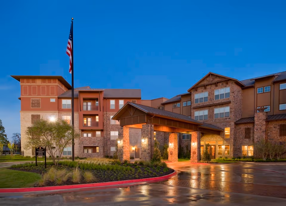 Exterior view of a multi-story senior living facility building at dusk with warm lights illuminating the entrance area, an American flag on a tall flagpole, and landscaped greenery around the driveway.